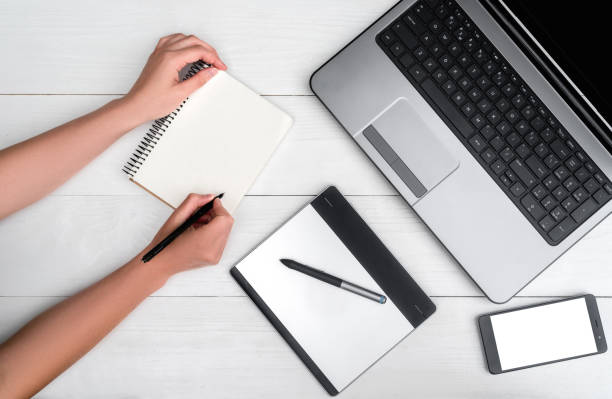 istock Top view on white wooden table with open laptop computer, cell phone, grafic tablet, empty diary and girl's hands writing in diary, free space. Mobile phone with white screen copy space 1322895690