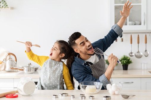 Cheerful arab father and little daughter having fun while baking together in kitchen, happy middle eastern dad and child singing and fooling, using spatula and whisk as microphones, copy space