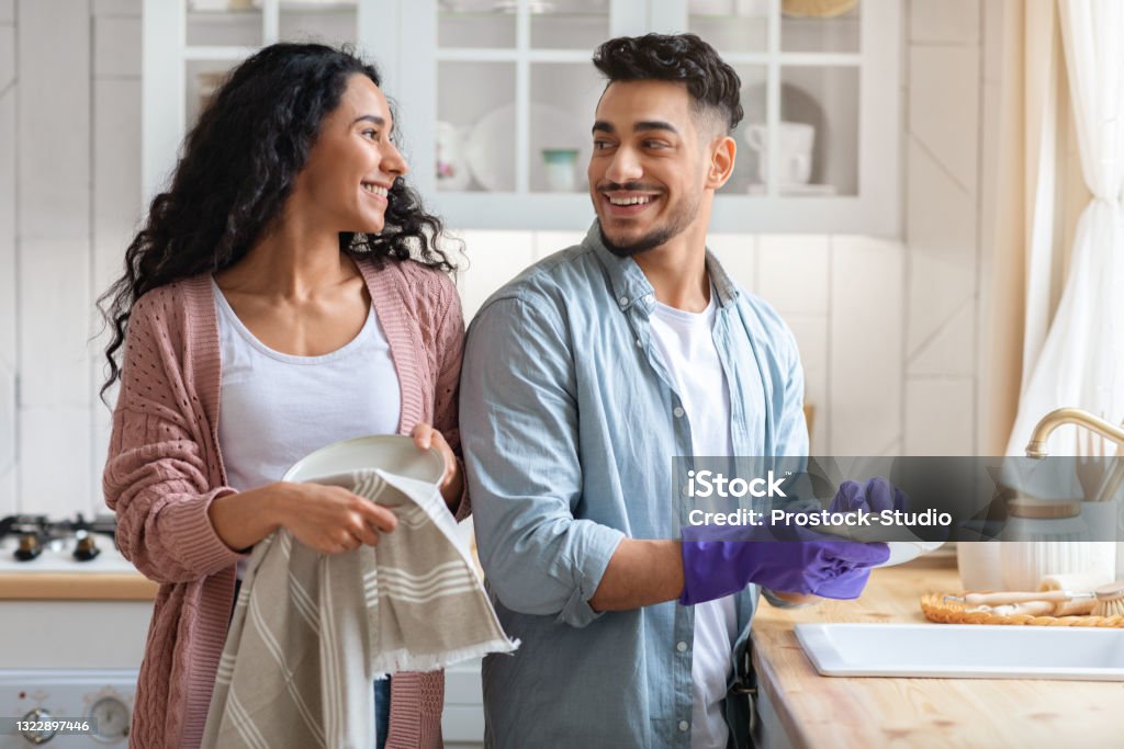 Cheerful Middle Eastern Couple Sharing Domestic Chores, Washing Dishes Together In Kitchen Cheerful Middle Eastern Couple Sharing Domestic Chores, Washing Dishes Together In Kitchen, Happy Millennial Arab Spouses Enjoying Making Cleaning At Home, Looking And Smiling To Each Other Couple - Relationship Stock Photo
