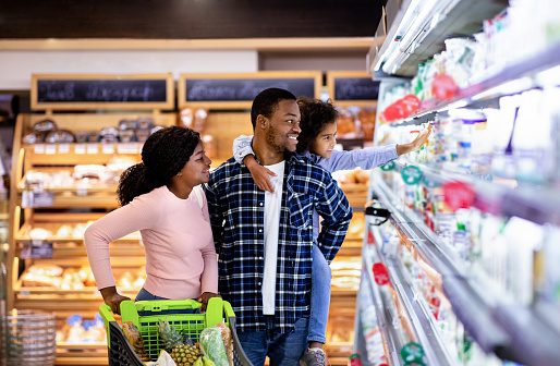Portrait of happy black family with trolley shopping together at grocery store. Millennial African American parents with lovely daughter picking food, choosing milk products at big mall