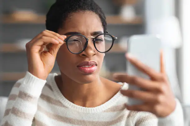 Photo of Astigmatism. Young Black Female In Eyeglasses Trying To Read Message On Smartphone