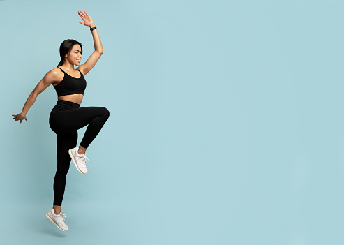 Fitness concept. Fit young african american woman jumping, lifting leg up, exercising and looking at empty space on blue background, studio shot, banner. Energy and workout