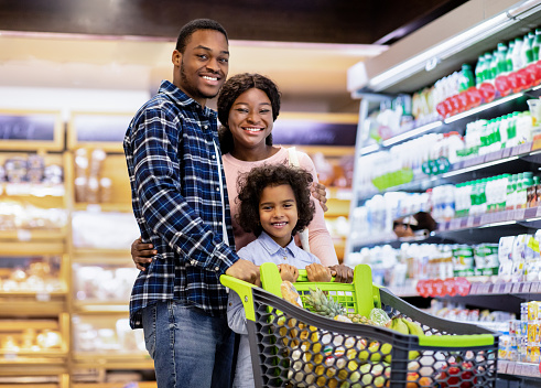 Portrait of joyful African American family with cute daughter posing and smiling at camera in big supermarket. Happy parents with child holding on to shopping cart full of food products at mall