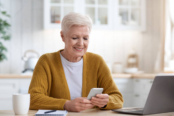 istock Smiling mature lady sitting at table with laptop, using smartphone 1322900043