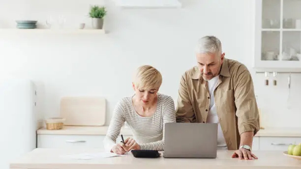 Photo of Concentrated senior man and woman counting expenses and pays financial bills with notepad and laptop