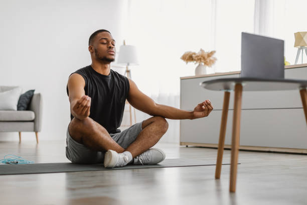 Peaceful African American Guy Meditating At Computer At Home Online Yoga. Peaceful African American Guy Meditating At Computer Sitting In Lotus Position In Front Of Laptop Watching Meditation Video Tutorial At Home. Meditation Relaxing Practice And Exercises meditation stock pictures, royalty-free photos & images