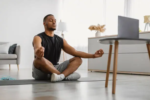 Online Yoga. Peaceful African American Guy Meditating At Computer Sitting In Lotus Position In Front Of Laptop Watching Meditation Video Tutorial At Home. Meditation Relaxing Practice And Exercises