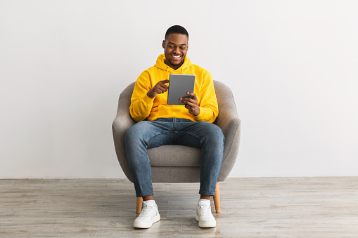 Happy Black Guy Using Digital Tablet Computer Browsing Internet Sitting In Chair Over Gray Wall Background. Online Technology And Modern Gadgets Lifestyle Concept