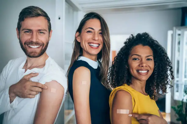 Three young friends pointing at their arms with a bandage after receiving COVID-19 vaccine. Young business people showing their shoulders after getting coronavirus vaccine.