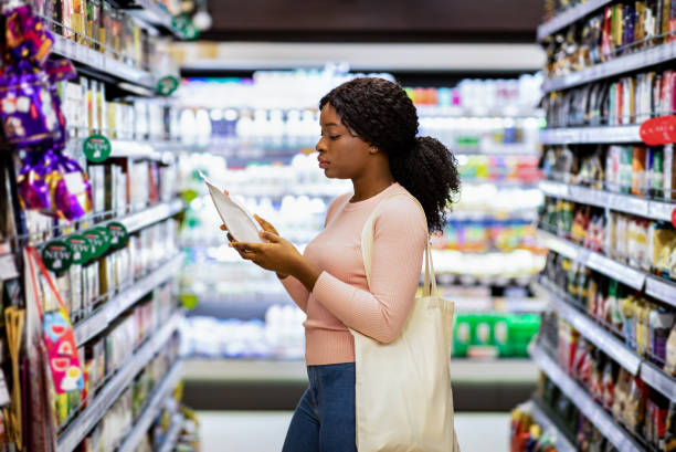 Pretty black lady with eco bag reading labels on food products, shopping for groceries at mall Pretty black lady with eco bag reading labels on food products, shopping for groceries at mall. Millennial African American woman making selection of groceries at modern supermarket reusable bag stock pictures, royalty-free photos & images