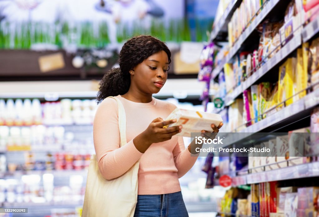 Attractive black woman with tote bag holding food product, buying groceries at supermarket Attractive black woman with tote bag holding food product, buying groceries at supermarket. Beautiful African American lady looking through labels at grocery department of huge mall Supermarket Stock Photo