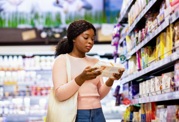 Attractive black woman with tote bag holding food product, buying groceries at supermarket Attractive black woman with tote bag holding food product, buying groceries at supermarket. Beautiful African American lady looking through labels at grocery department of huge mall supermarket stock pictures, royalty-free photos & images