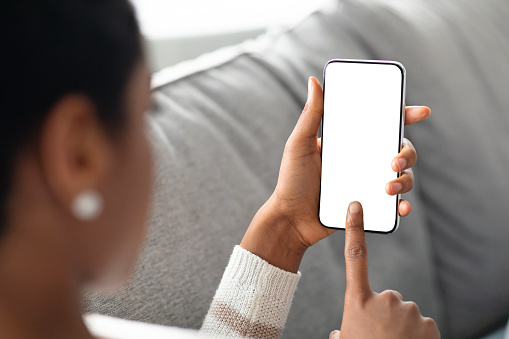 Unrecognizable Black Female Touching Blank Smartphone Touchscreen At Home, Over Shoulder View Of African Woman Browsing New App Or Website On Modern Gadget Mockup Image With Copy Space, Closeup