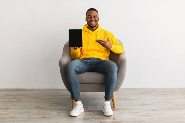 istock African Guy Showing Tablet Screen Sitting In Chair, Gray Background 1322904665