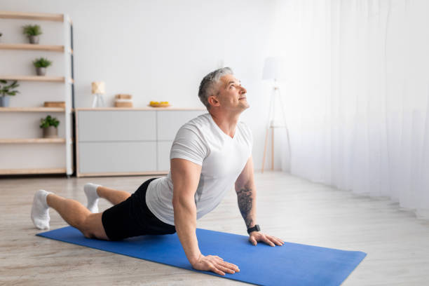 aktiver senior mann tun kobra pose auf yogamatte, training im wohnzimmer innenraum, leeren raum - core strength stock-fotos und bilder