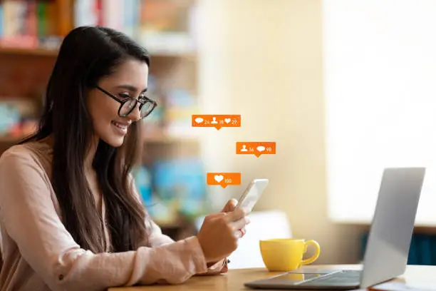 Happy latin female blogger using smartphone with social media notification icons, sitting in cafe, free space. Phone user networking online, using social media and reading feed news