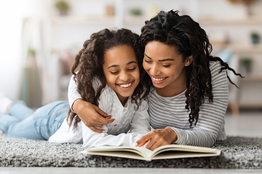 Pretty black girl reading book with her mom, laying together on floor and hugging, home interior. Happy african american mother and daughter reading books and embracing, closeup