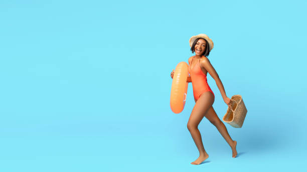 istock Hello summer. Positive black lady in bikini doing to beach with straw bag and inflatable ring on blue studio background 1322908409