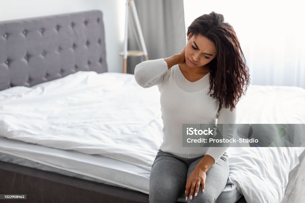 Portrait of black woman with neck pain sitting on bed Exhausted Person. Tired African American lady sitting on bed massaging sore neck, feeling pain after sleeping on bed uncomfortable mattress, free copy space. Black woman suffering from inflammation Fibromyalgia Stock Photo