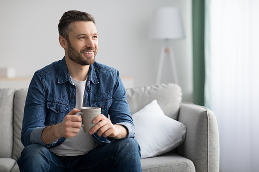 Cheerful bearded man enjoying cup of coffee at home, sitting on couch in living room and looking at copy space. Handsome middle-aged man with mug of tea chilling alone, having good time
