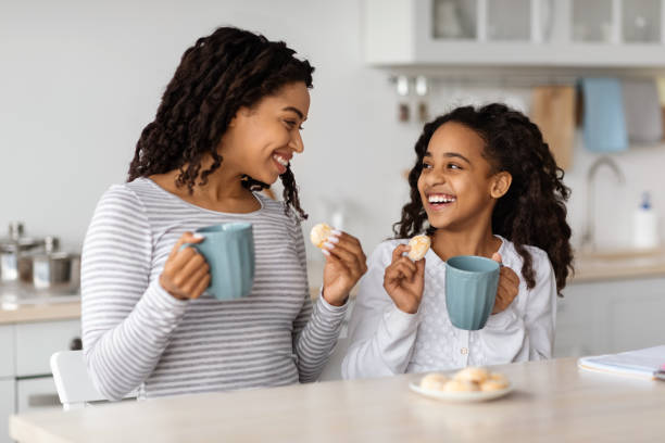Cute black mother and daughter drinking tea with cookies at kitchen, looking at each other and smiling, copy space. Happy african american mummy and kid having snack together at home