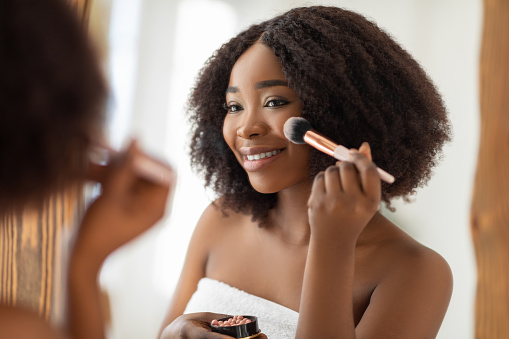 Natural makeup for young skin. Sexy black lady putting on blusher or powder with brush in front of mirror indoors. Sensual African American woman applying organic decorative makeup