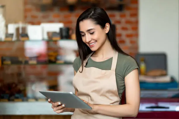 Young female owner of modern cafe in apron holding digital tablet and standing with shopwindow on background, using pad, copy space. Modern technology and work in cafeteri