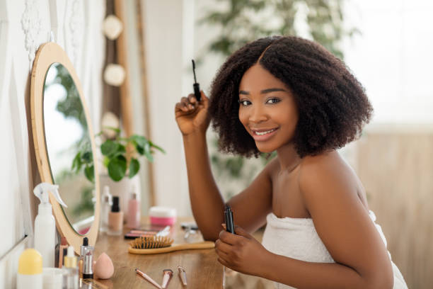 istock Morning beauty routine. Attractive black lady applying mascara, sitting at dressing table, looking in mirror at home 1322910042