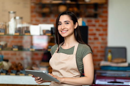 Apertura de pequeñas empresas. Mujer árabe feliz en delantal cerca del mostrador del bar sosteniendo la tableta digital y mirando la cámara photo