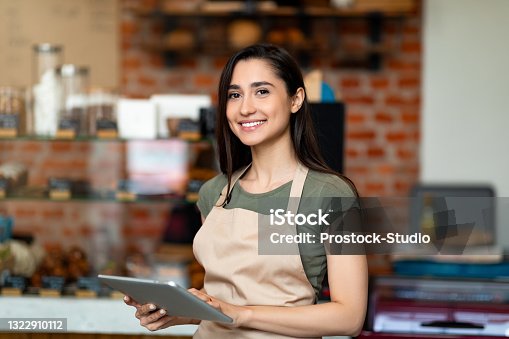 istock Opening small business. Happy arab woman in apron near bar counter holding digital tablet and looking at camera 1322910112