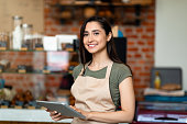 Opening small business. Happy arab woman in apron near bar counter holding digital tablet and looking at camera