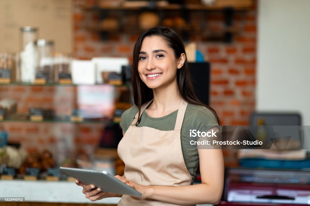 Opening small business. Happy arab woman in apron near bar counter holding digital tablet and looking at camera Opening small business. Happy arab woman in apron near bar counter holding digital tablet and looking at camera, waiting for clients in modern loft cafe Entrepreneur Stock Photo