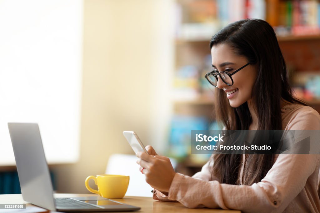 Freelance concept. Happy arab woman working on pc at cafe, drinking tea and using smartphone, having break Freelance concept. Happy arab woman working on pc at cafe, drinking tea and using smartphone, having break during distant work, empty space. Modern lifestyle concept Businesswoman Stock Photo