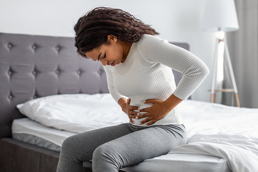 Portrair of upset black woman suffering from strong abdominal pain, touching her tummy, sitting on bed. Sad young lady feeling acute stomachache, free copy space. Chronic disease, spasm, hernia