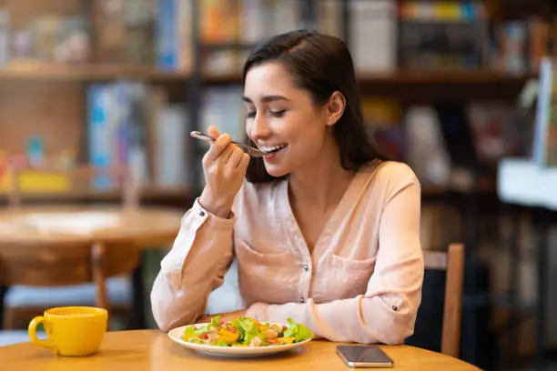 Happy latin woman eating lunch in cafe, enjoying delicious salad with closed eyes and drinking hot beverage. Lady eating healthy meal, sitting indoors in restaurant, free space