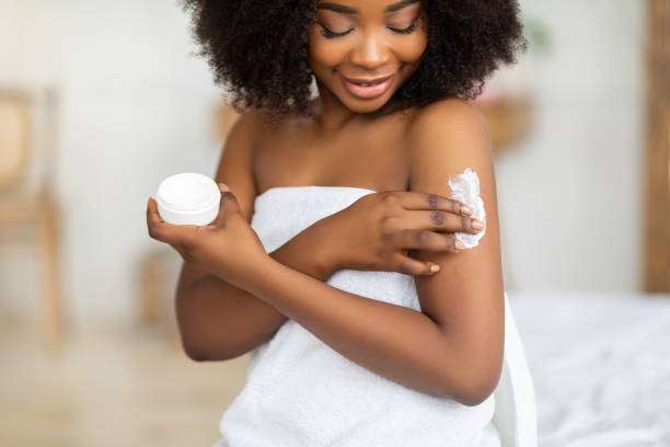 beautiful black lady wearing towel after bath, applying cream, rubbing in nourishing body butter at home - moisturizer women cosmetics body imagens e fotografias de stock