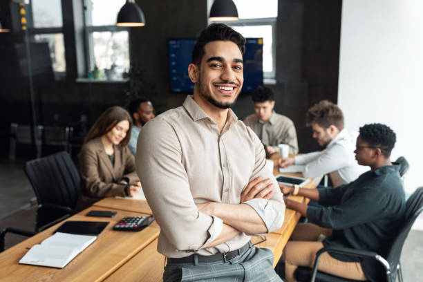 Young bearded businessman sitting on desk and posing Successful Person. Portrait of confident smiling bearded businessman sitting leaning on desk in office, posing with folded arms and looking at camera, colleagues working in blurred background bossy stock pictures, royalty-free photos & images