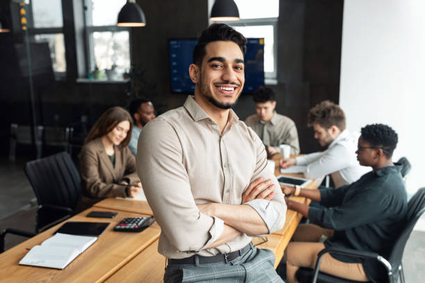 Young bearded businessman sitting on desk and posing Successful Person. Portrait of confident smiling bearded businessman sitting leaning on desk in office, posing with folded arms and looking at camera, colleagues working in blurred background confidence stock pictures, royalty-free photos & images