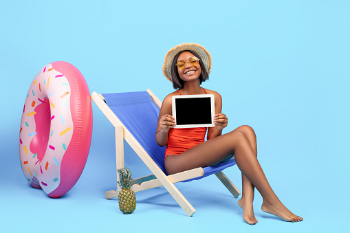 Positive black woman in swimsuit holding tablet with empty screen while relaxing in lounge chair on blue studio background, mockup for your application or website design