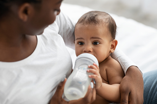 Childcare Concept. Closeup portrait of happy African American woman holding baby bottle and feeding her cute little black with milk at home. Nutrition, Love, Care And Family, Babysitting