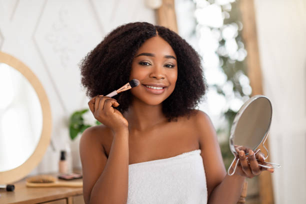 istock Smiling black woman applying cosmetic powder or blusher on her face, using decorative makeup, looking in mirror at home 1322913910