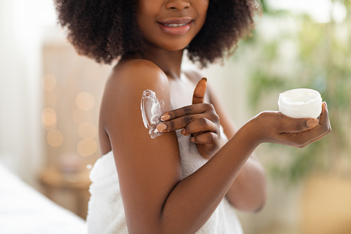 Cropped view of young black woman applying cream on her body, pampering her skin after shower at home, closeup. Unrecognizable African American lady making daily beauty routine