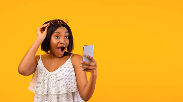 istock Cool mobile application. Shocked black woman using smartphone, opening mouth in excitement, orange background 1322914056