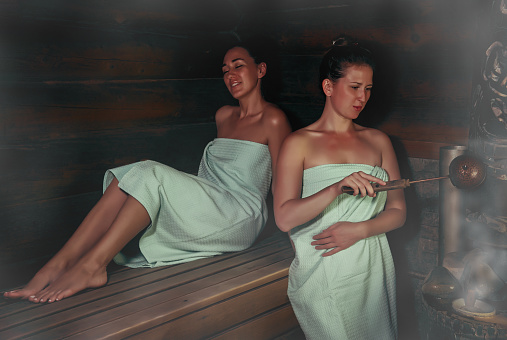 two women in a traditional steam sauna banya pours water on hot stones on stove