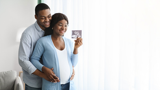Attractive african american couple posing with baby sonogram, home interior, panorama with copy space. Happy black husband hugging his wife, showing ultrasound image of their kid. Parenthood concept