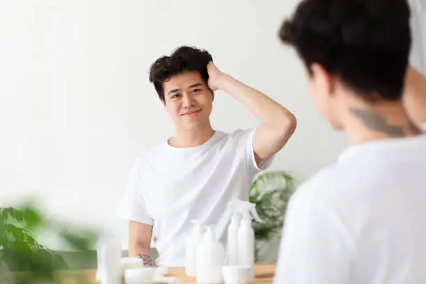 Modern cosmetics for male beauty, daily procedures and care. Smiling asian millennial guy touches through thick hair, enjoys and looks at reflection in mirror in bathroom interior with bottles