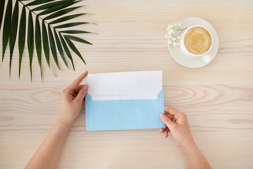 Top view of female hands opening greeting card with blue envelope on wooden table with cup of coffee and tropical leaf, flat lay. Mockup. Minimalistic woman's desk