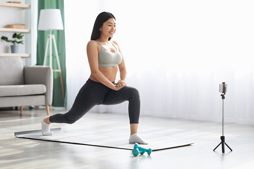 Smiling Asian Woman Training At Home In Front Of Smartphone On Tripod, Beautiful Korean Female Fitness Blogger Shooting Online Video Fitness Tutorials, Showing Stretching Exercises At Camera