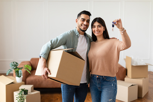 House Ownership. Young Couple Showing Keys And Holding Cardboard Box, Cheerful Guy And Lady Hugging After Moving In New Apartment Standing In Living Room. Insurance, Real Estate, Mortgage Concept