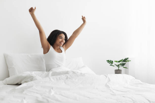 relaxado depois de boa noite de sono. feliz mulher afro-americana sentada na cama com cobertor, esticando depois de acordar - wake up stretching women black - fotografias e filmes do acervo